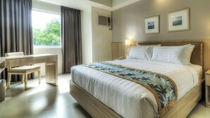 Hotellihuone, jossa on suuri sänky, työpöytä, tuoli, ikkuna verhoilla sekä kaksi kehystettyä kuvaa seinällä.