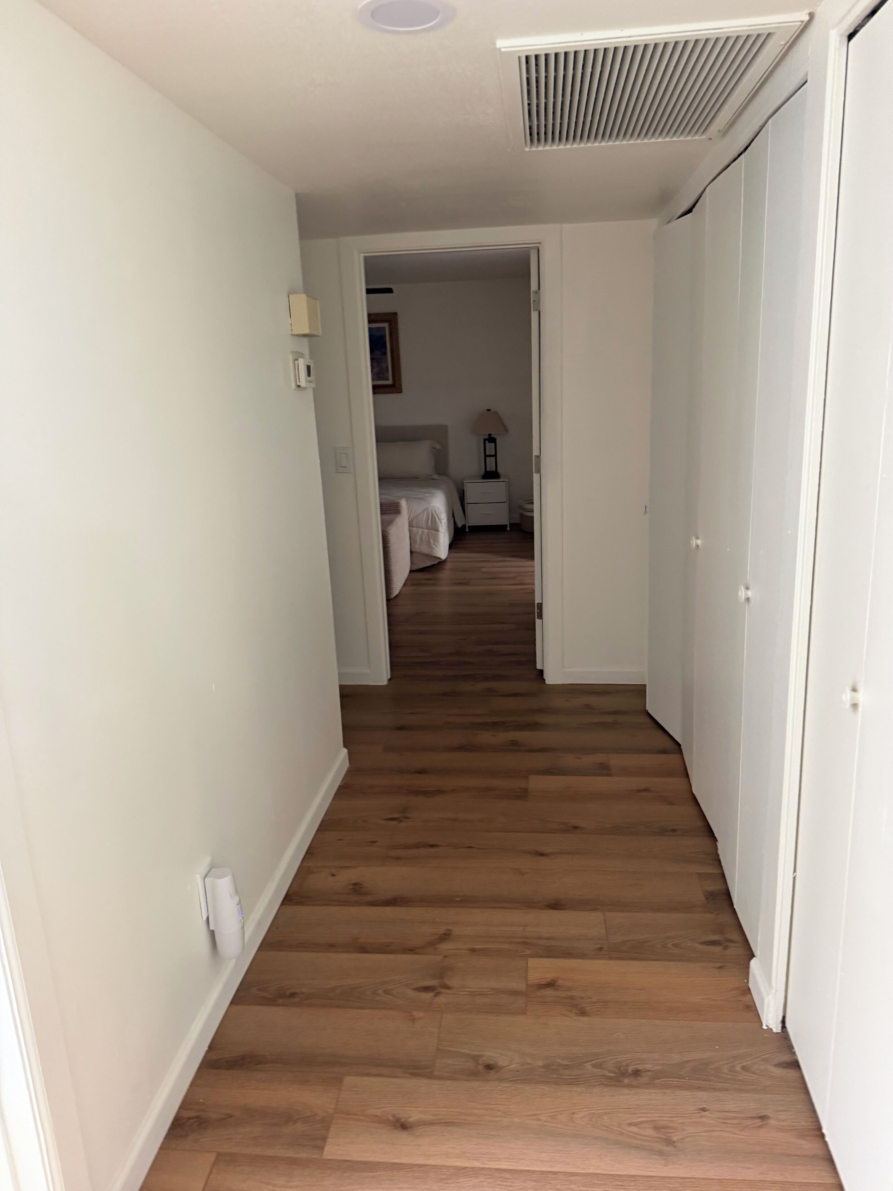 Un pasillo con piso de madera que lleva a un dormitorio con cama y mesita de noche.