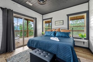 Ein Schlafzimmer mit einem großen Bett, einer Holzbalkendecke, Blick durch eine Schiebetür und einem Fenster mit Jalousien.