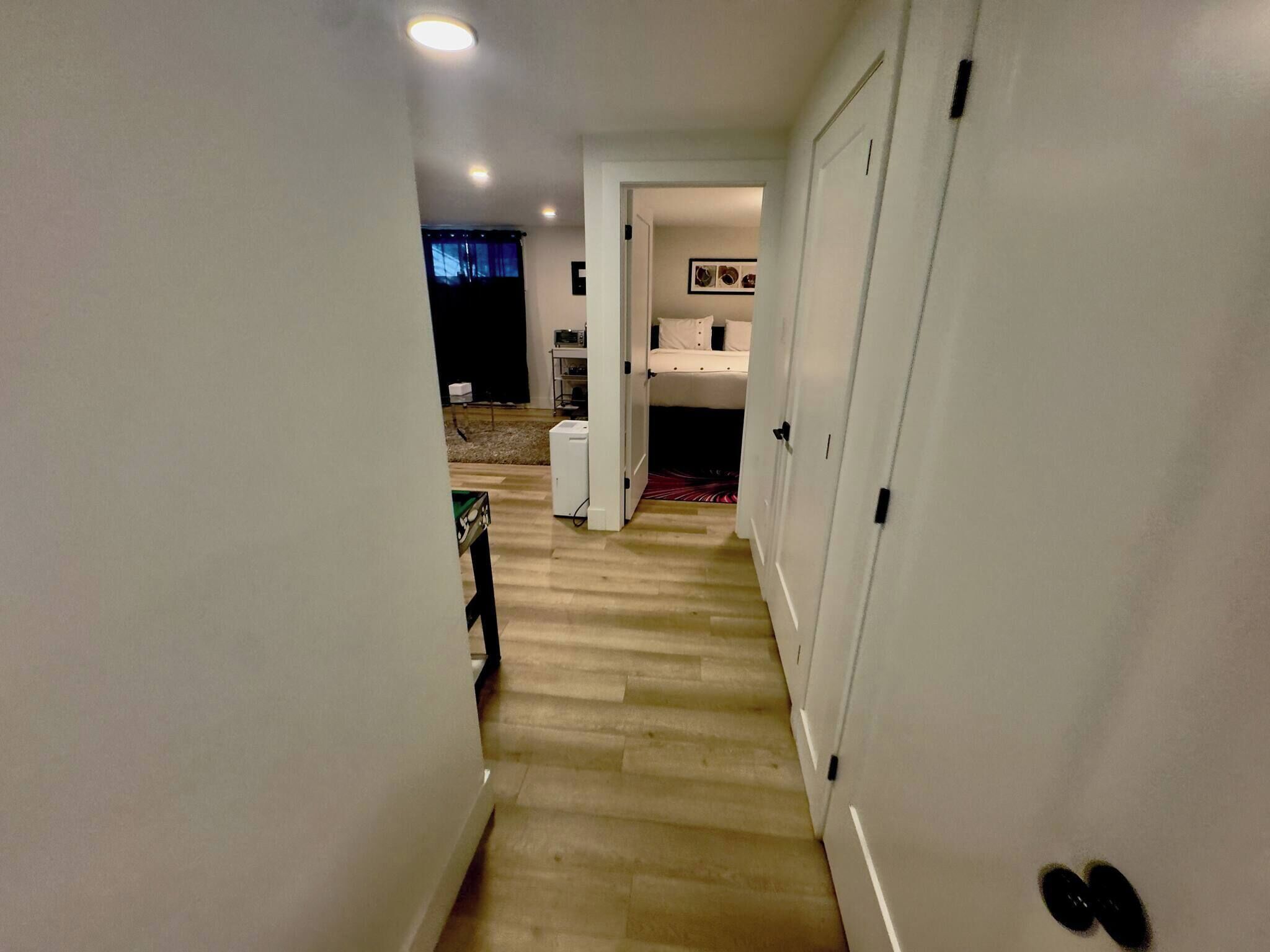 Un pasillo estrecho con piso de madera, paredes blancas y una puerta que da a una habitación con cama y una mesita.
