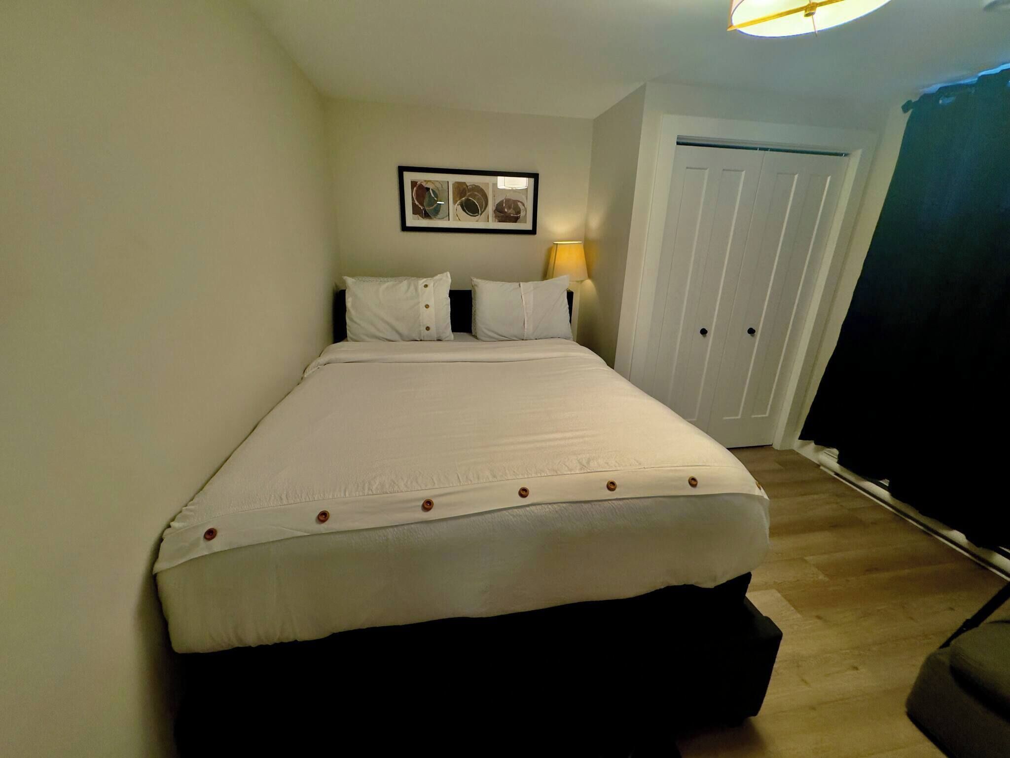 A neatly made bed with a white duvet, a framed picture above it, and a closed door to the right.