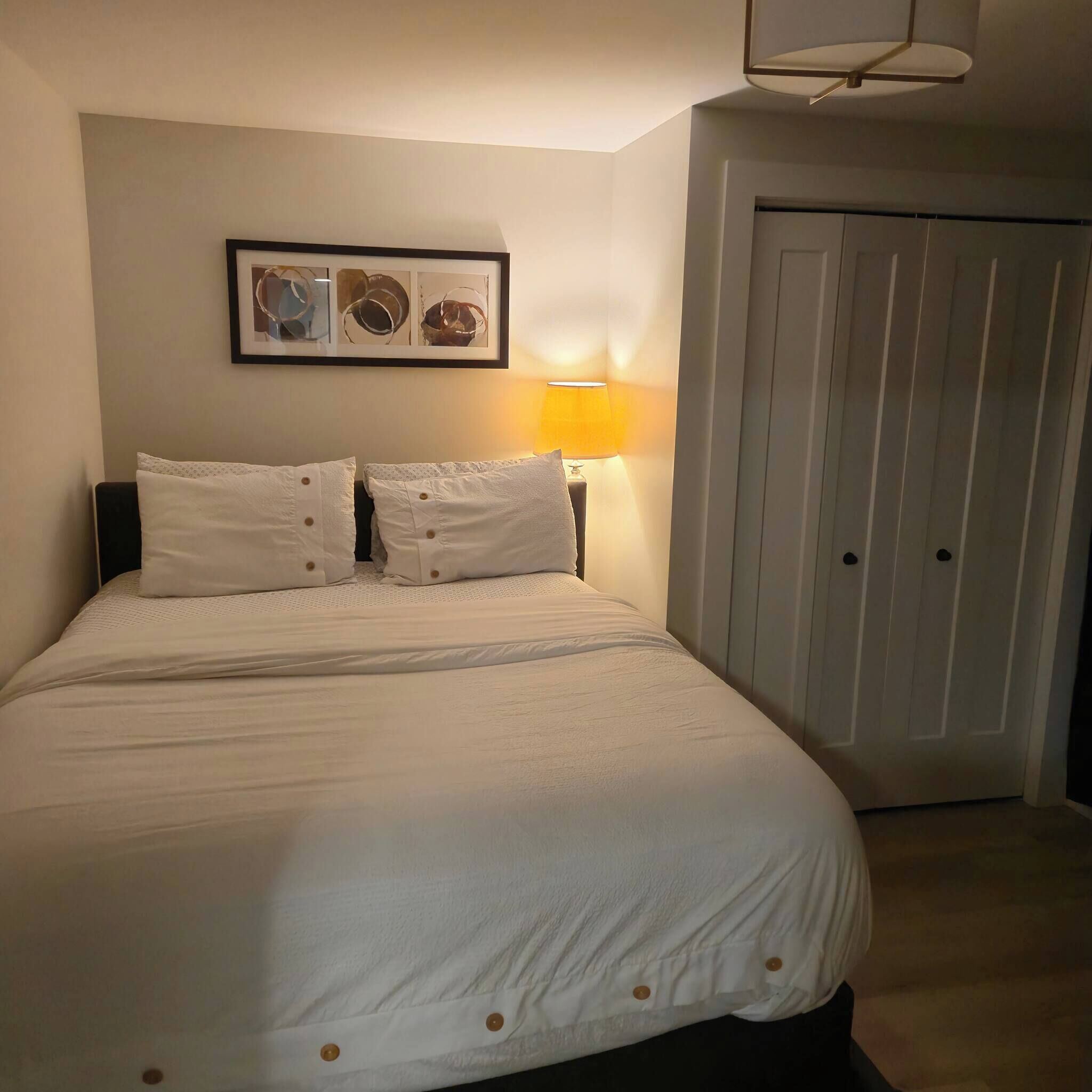 Una cama bien tendida con sábanas blancas y botones, una obra de arte abstracta enmarcada encima, y una lámpara a la derecha.