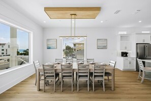 A modern dining area with a wooden table and chairs, a view of the city through large windows, and a kitchen with white cabinets and stainless steel appliances. - Brand New Luxe Home for Family Gatherings • Elevator • Gulf Views + Heated Pool (Panama City Beach)