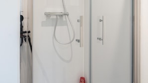 Um chuveiro com divisória de vidro, um chuveiro manual e um painel de controle fixado na parede, além de uma garrafa vermelha na parede.