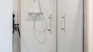 A glass-enclosed shower with a handheld showerhead, a wall-mounted control panel, and a red bottle on the wall.