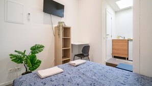 Quarto com cama, escrivaninha, cadeira, televisão, planta e banheiro visível através de uma porta aberta.