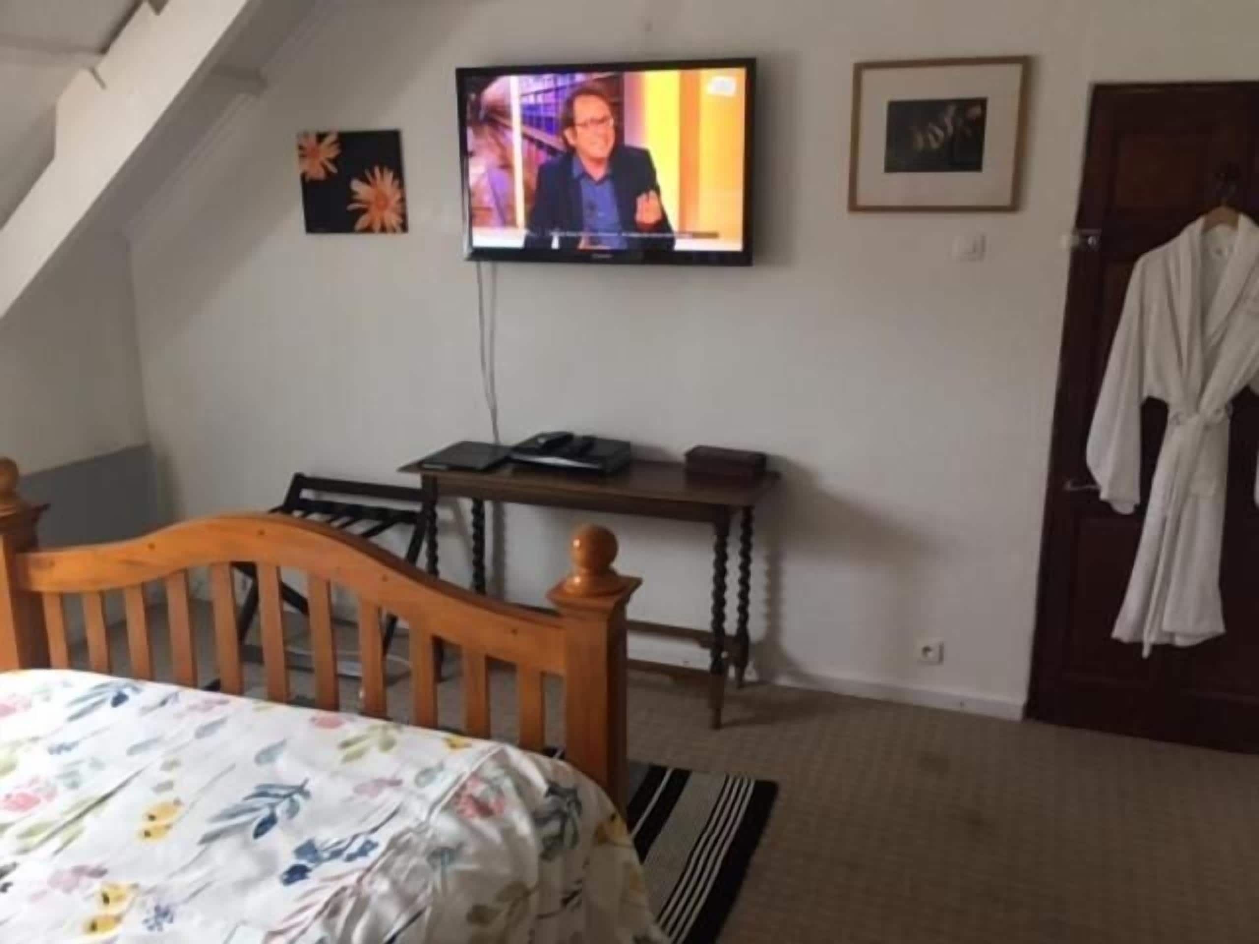 Un dormitorio con una cama, un televisor montado en la pared, una silla de madera, una mesita y una bata colgada en la puerta.