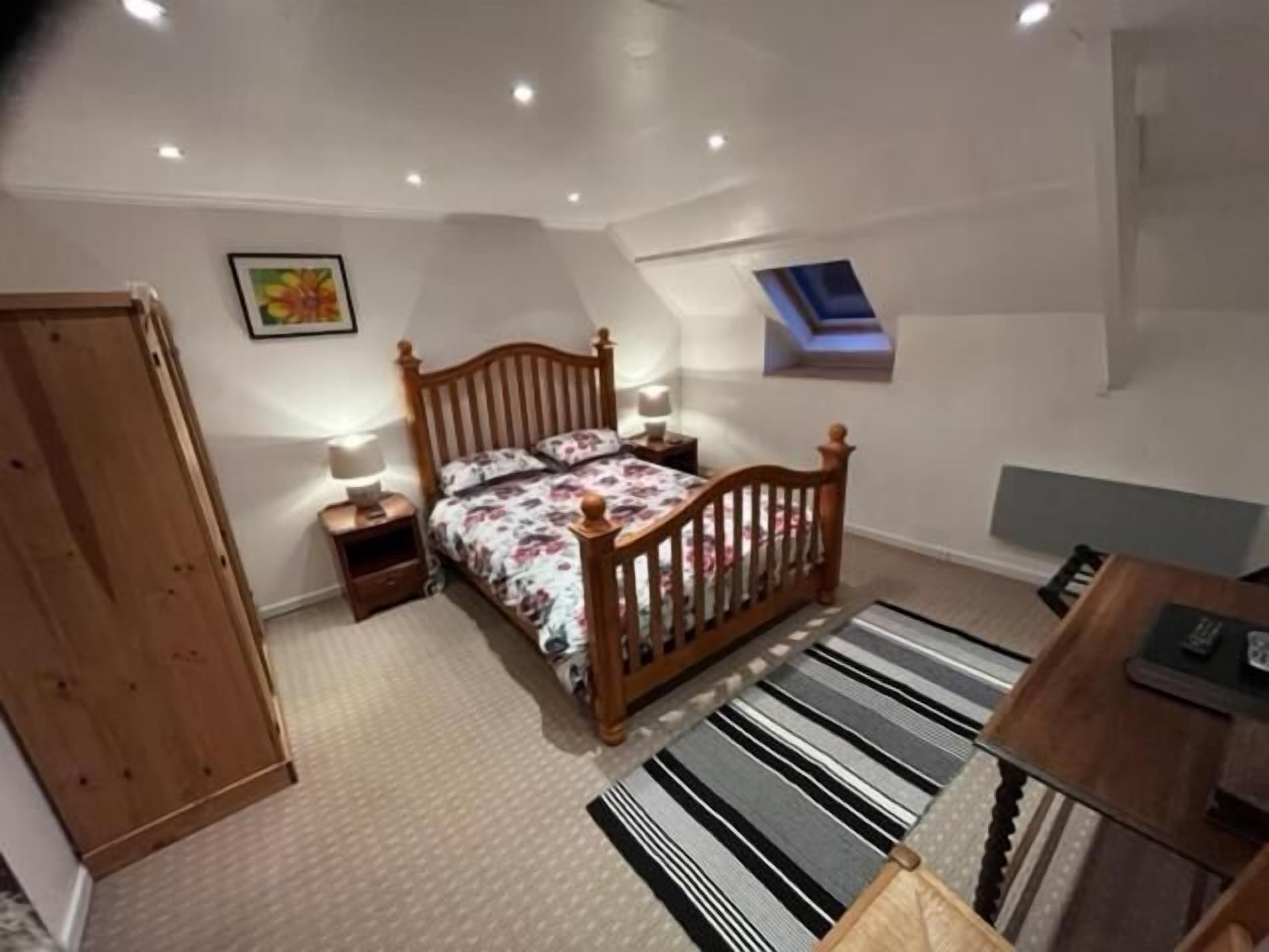 Une chambre à coucher avec un lit en bois, une table de chevet, un bureau, une chaise, une armoire, une fenêtre et un luminaire au plafond.