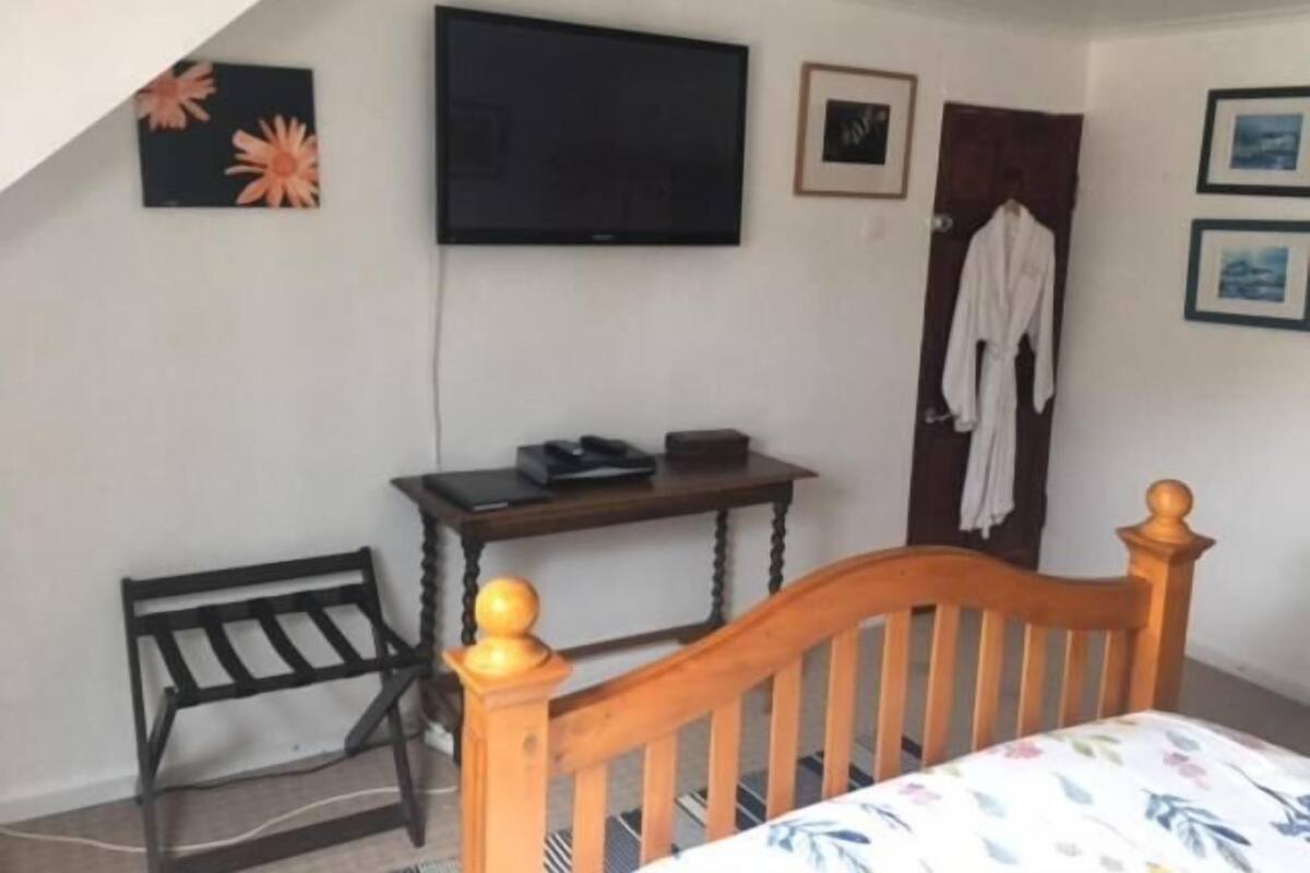 Un dormitorio con cama, televisor montado en la pared, una silla de madera, una mesita pequeña y una puerta con una bata colgada.