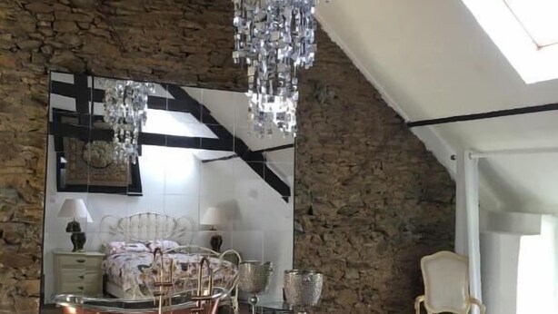 A room with a stone wall, a large mirror, a bed, a chandelier, and copper bathtub.