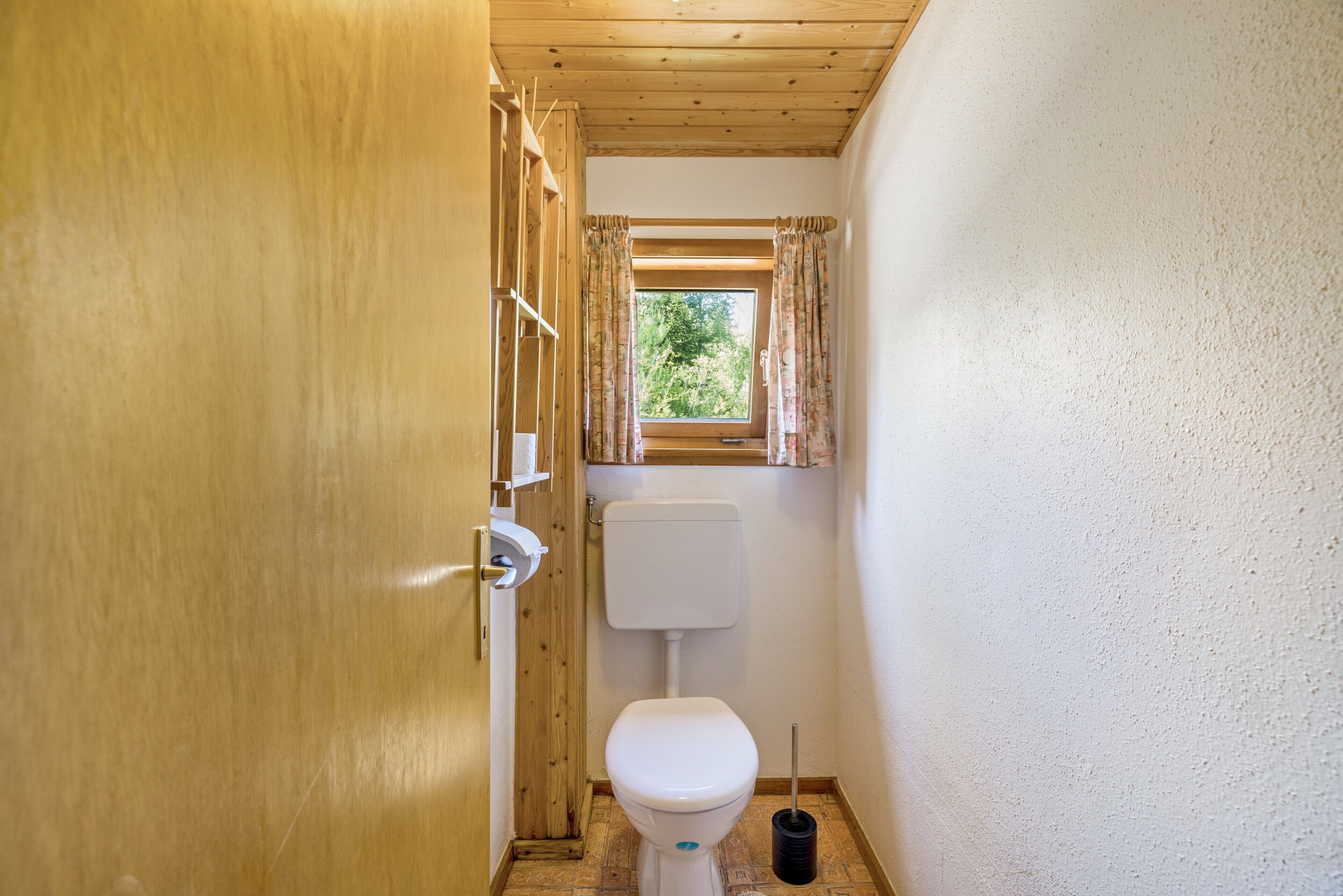 Une petite salle de bain avec une porte en bois, des toilettes, une fenêtre avec des rideaux et une petite étagère contenant des articles de toilette.