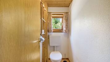 Une petite salle de bain avec une porte en bois, des toilettes, une fenêtre avec des rideaux et une petite étagère contenant des articles de toilette.
