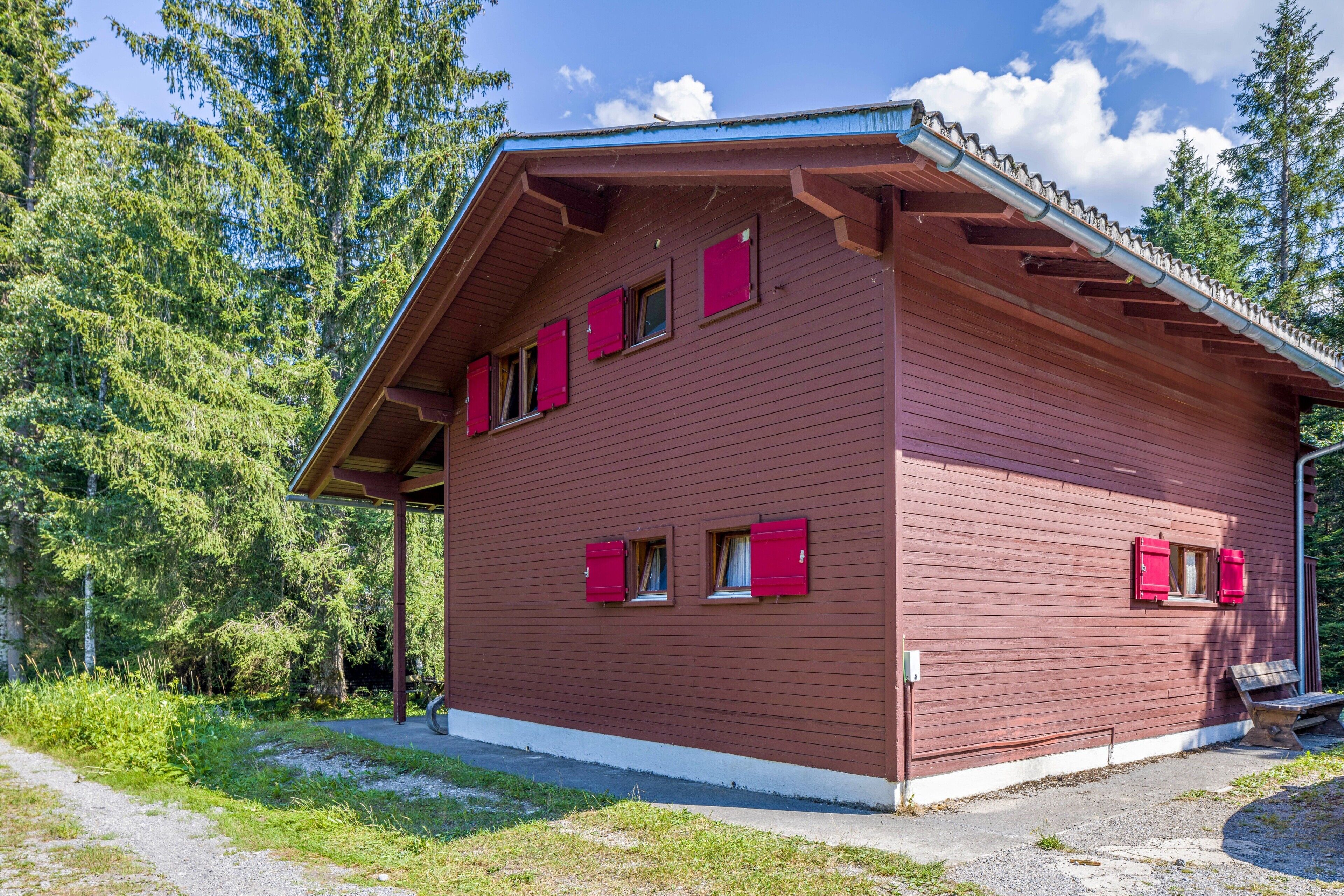 Punaisilla ikkunaluukuilla varustettu, kaksikerroksinen puinen mökki, jonka ympärillä on puita.