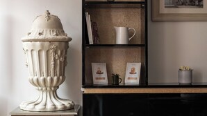 Um quarto com um vaso branco sobre uma mesinha, uma estante com livros e um quadro emoldurado, e outro quadro na parede.