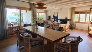 Een ruime kamer met een houten eettafel, omringd door rieten stoelen, een bank en een open haard.