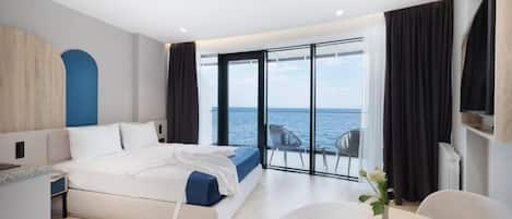 A modern hotel room with a large bed, a dining area, and a view of the sea.