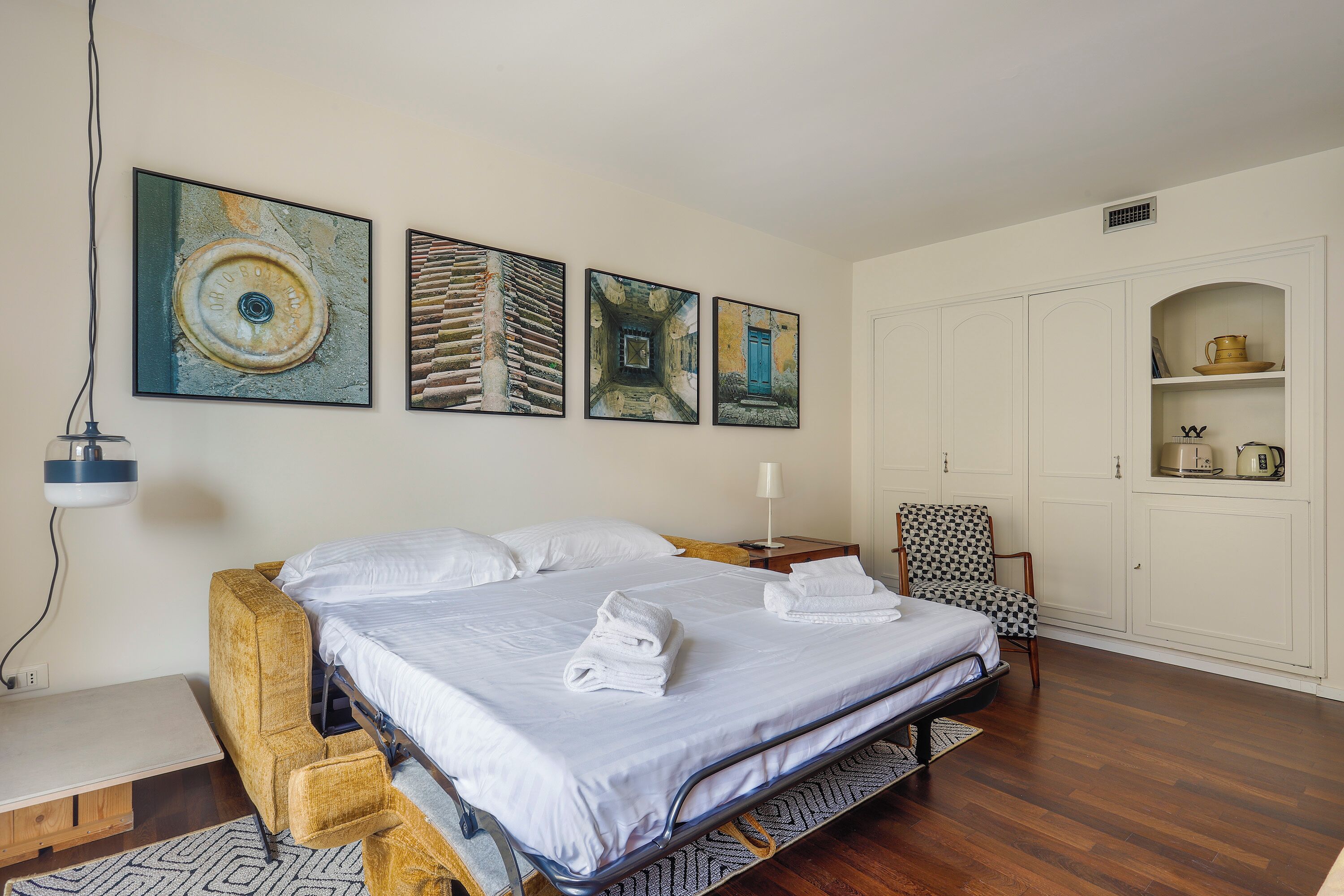 A hotel room with a bed, artwork on the wall, a chair, and a cabinet with items on it.