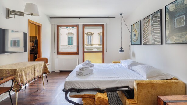 Hotellihuone, jossa on sänky, pöytä pöytäliinalla, tuoli, sohva ja televisio.