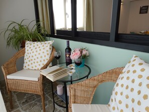 Una silla de mimbre con un cojín de lunares, una mesa de vidrio con un libro, vino y flores, además de una planta en maceta.