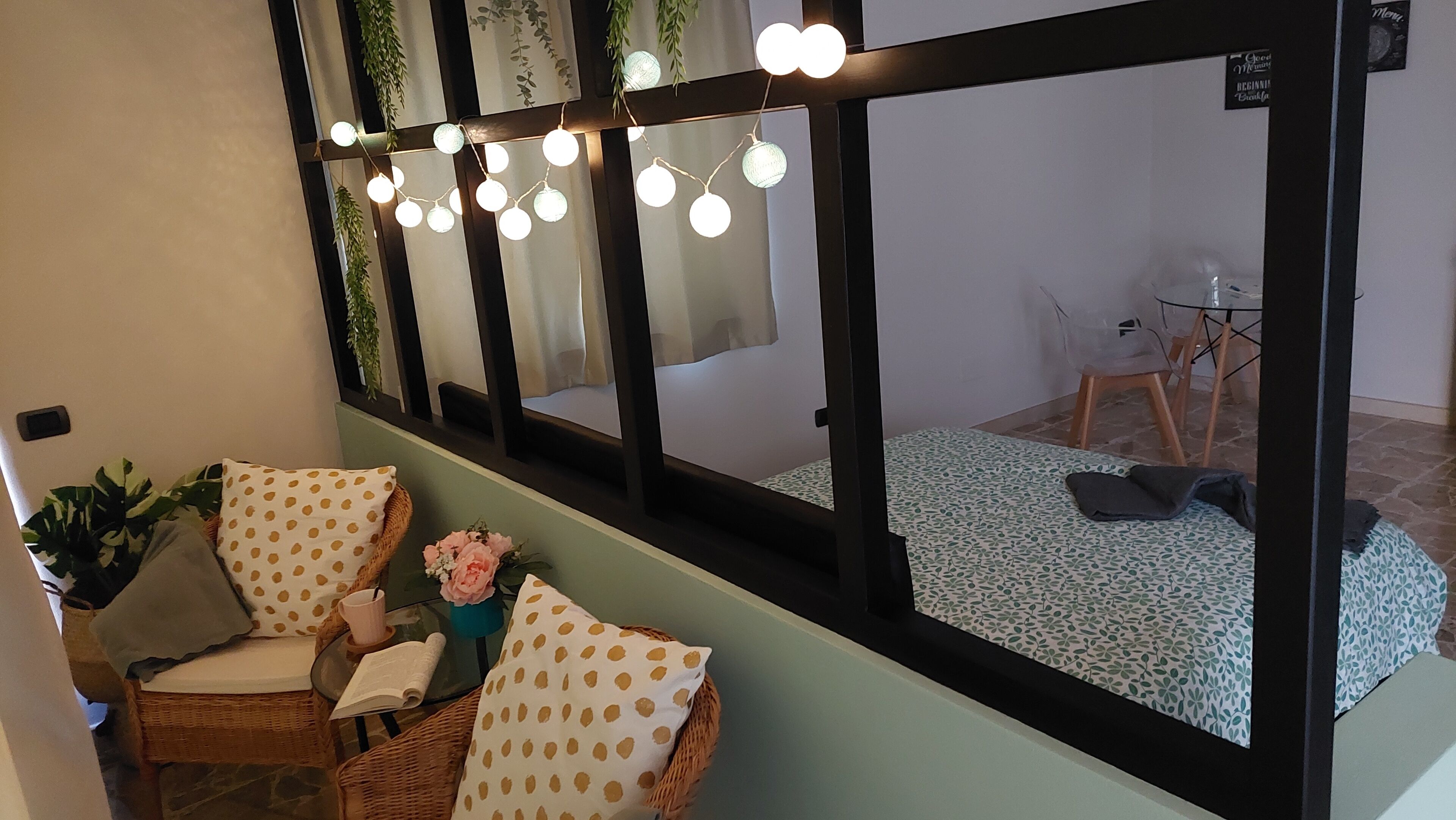 A room with a large mirror, a wicker chair, a small table with a book, and a bed with patterned bedding.