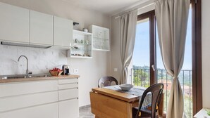 A kitchen with white cabinets, a wooden table, and a view of the outdoors through a window with curtains. - Apartment 'A Fosdinovo' with Mountain View, Wi-Fi and Air Conditioning (Fosdinovo)