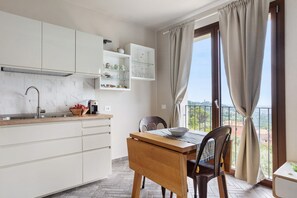 A kitchen with white cabinets, a wooden table, and a view of the outdoors through a window with curtains. - Apartment 'A Fosdinovo' with Mountain View, Wi-Fi and Air Conditioning (Fosdinovo)