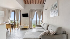 A modern living room with a sofa, dining table, and chairs, large windows with curtains, and a mounted artwork. - Apartment 'A Fosdinovo' with Mountain View, Wi-Fi and Air Conditioning (Fosdinovo)