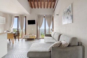 Una sala de estar moderna con sofá, mesa de comedor y sillas, ventanas amplias con cortinas, y una obra de arte colgada.