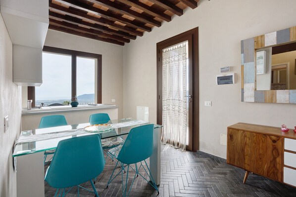A dining area with a glass table, teal chairs, a wooden cabinet, and a view through a window. - Apartment 'Incantevole - Fosdinovo' with Mountain View, Wi-Fi and Air Conditioning (Fosdinovo)