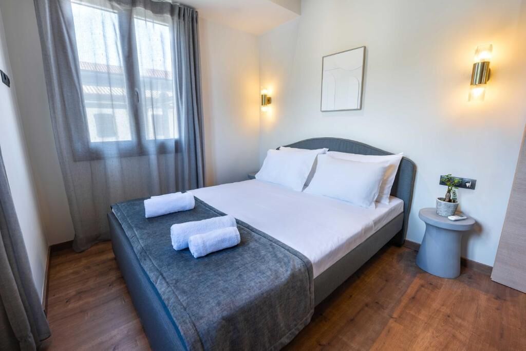 Una cama bien tendida con sábanas blancas y dos toallas, una mesita de noche con una planta, y una lámpara fijada a la pared.