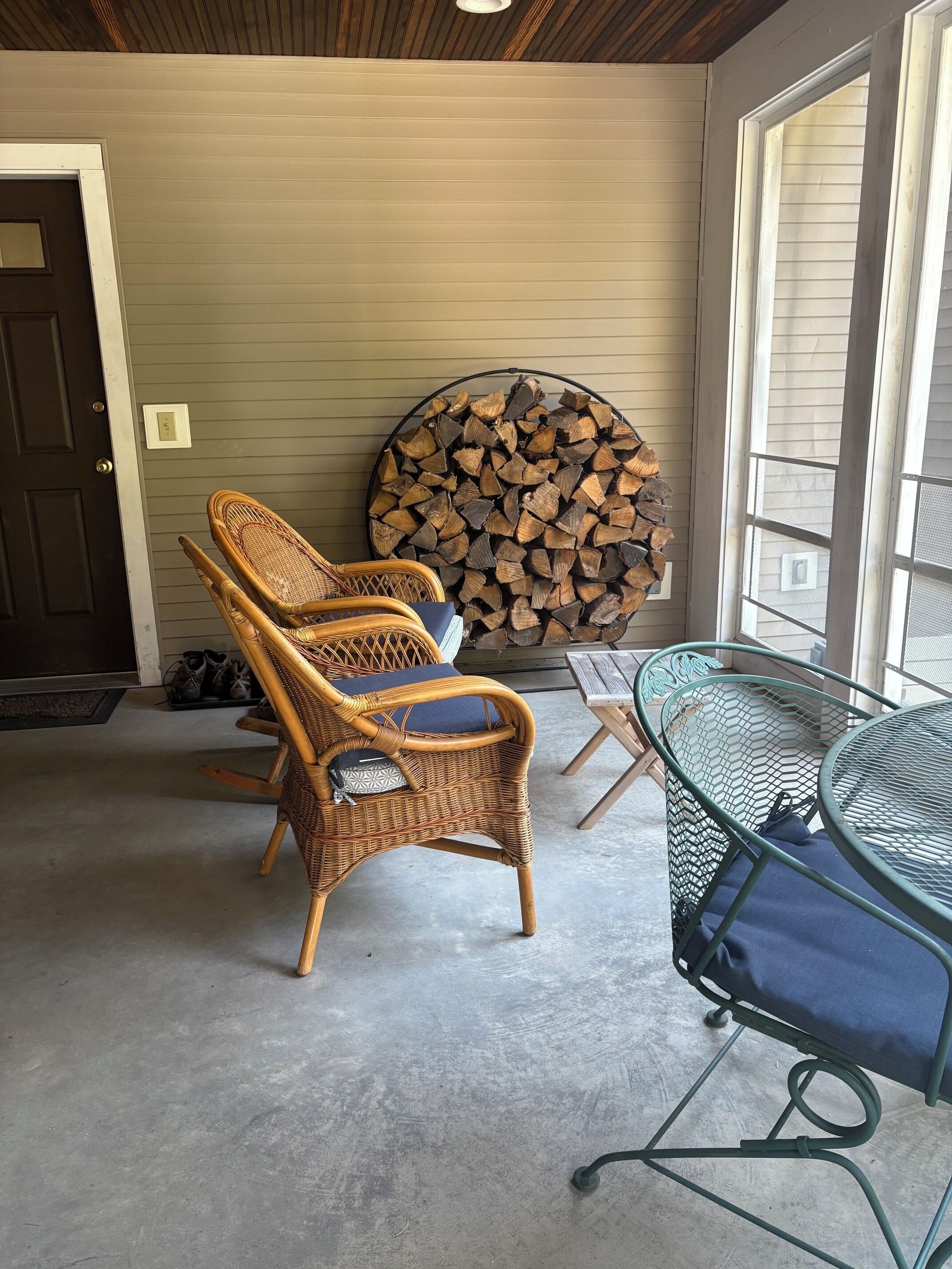A room with wicker chairs, a metal table, a stack of firewood, and a door.