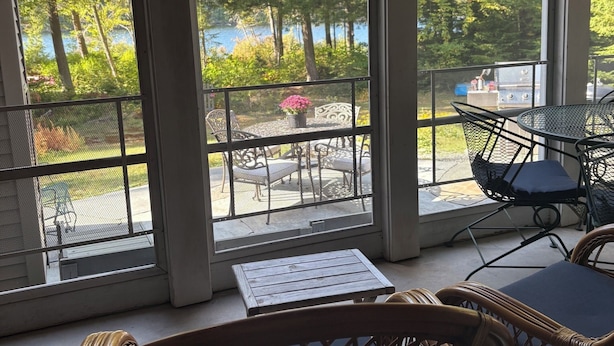 Eine überdachte Terrasse mit Korbmöbeln, einem kleinen Tisch und Blick auf einen See und Bäume.