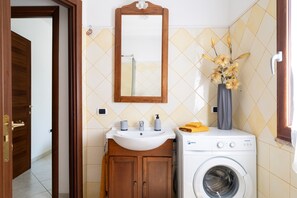 Casa de banho com porta de madeira, pia branca, espelho, máquina de lavar roupa e um vaso com flores secas.