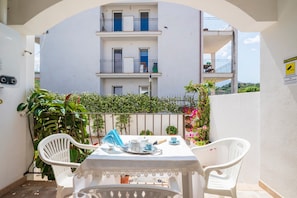 Varanda com mesa posta para dois, uma cadeira branca e vista de um edifício com várias varandas.