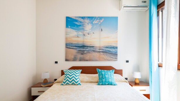 Une chambre à coucher comprenant un lit, deux tables de chevet, un climatiseur fixé au mur et un grand tableau représentant un paysage marin.