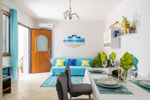 Zona pranzo con tavolo imbandito per quattro persone, divano blu con cuscini decorati, lampadario e mensola a parete con oggetti ornamentali.