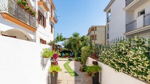 Une ruelle étroite, bordée de plantes en pot et d’une pelouse verdoyante, et flanquée d’immeubles blancs dotés de balcons.