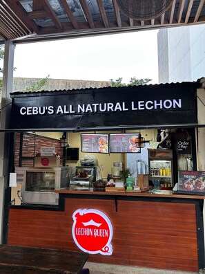 Un puesto de comida llamado 'CEBU'S ALL NATURAL LECHON', con un tablero de menú, una caja registradora y un letrero que dice 'LECHON QUEEN'.