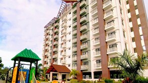 Un parque infantil con un sube y baja y una estructura para escalar, rodeado de un césped cuidado, con un edificio residencial de varios pisos al fondo.