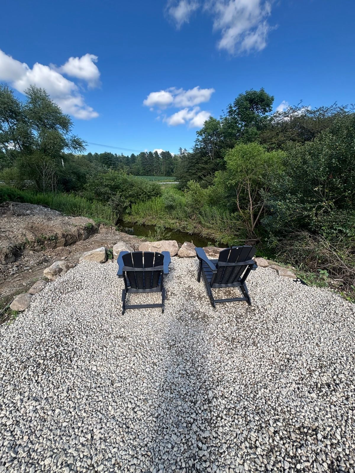 Two Adirondack chairs on a gravel surface with a natural landscape in the background.