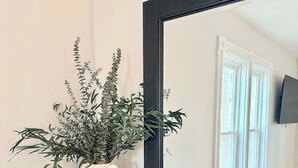 Un miroir avec un cadre noir, un vase blanc contenant une plante et des objets décoratifs posés sur une commode en bois foncé.