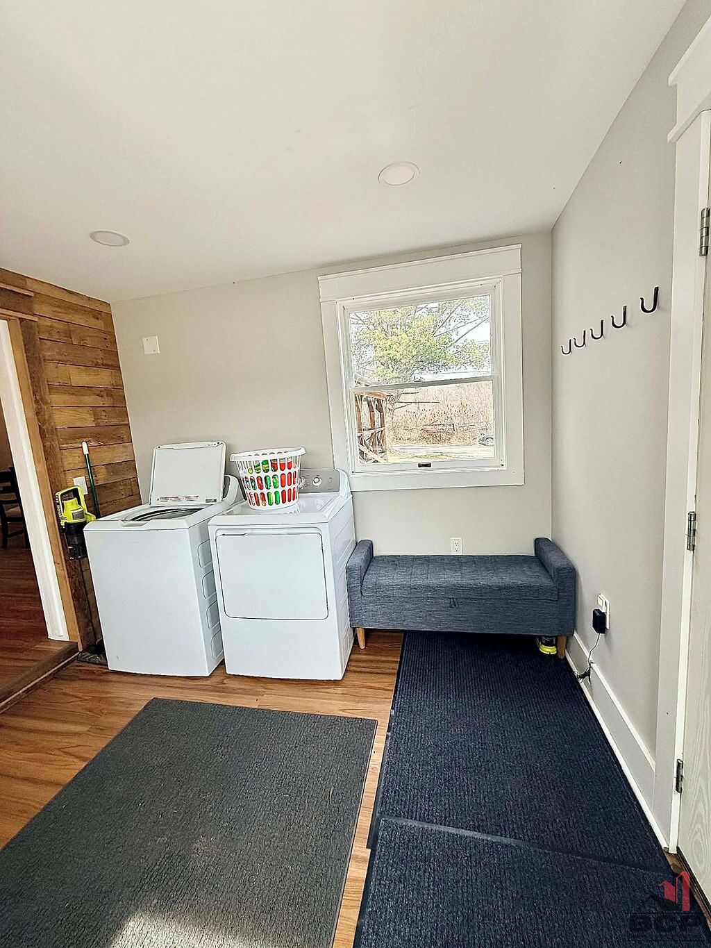 A laundry room with two white washing machines, a gray sofa, a window with a view, and a door with a sign.