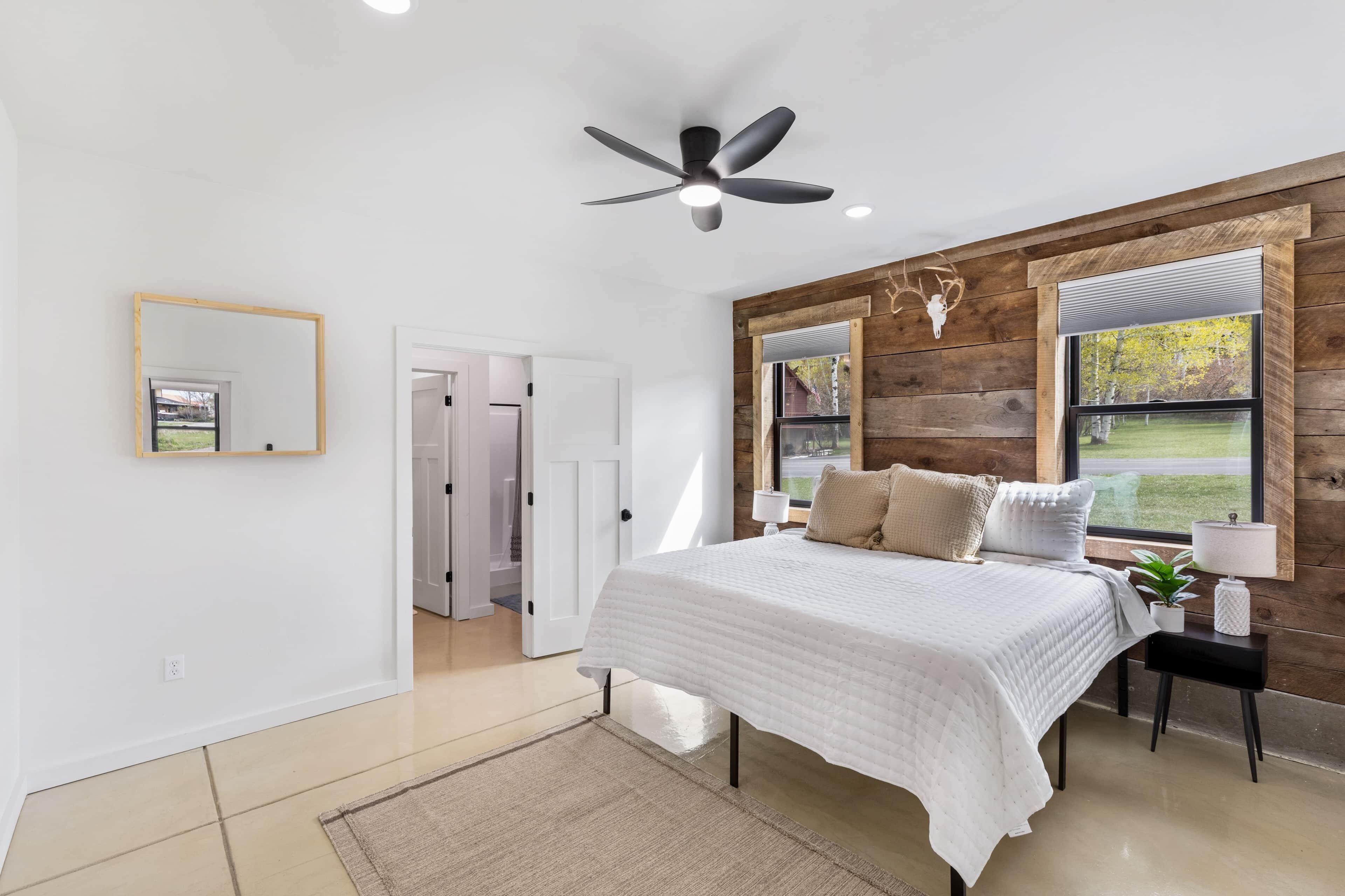 Un dormitorio con cama, ventilador de techo, ventana con vistas, pared de madera y una puerta.