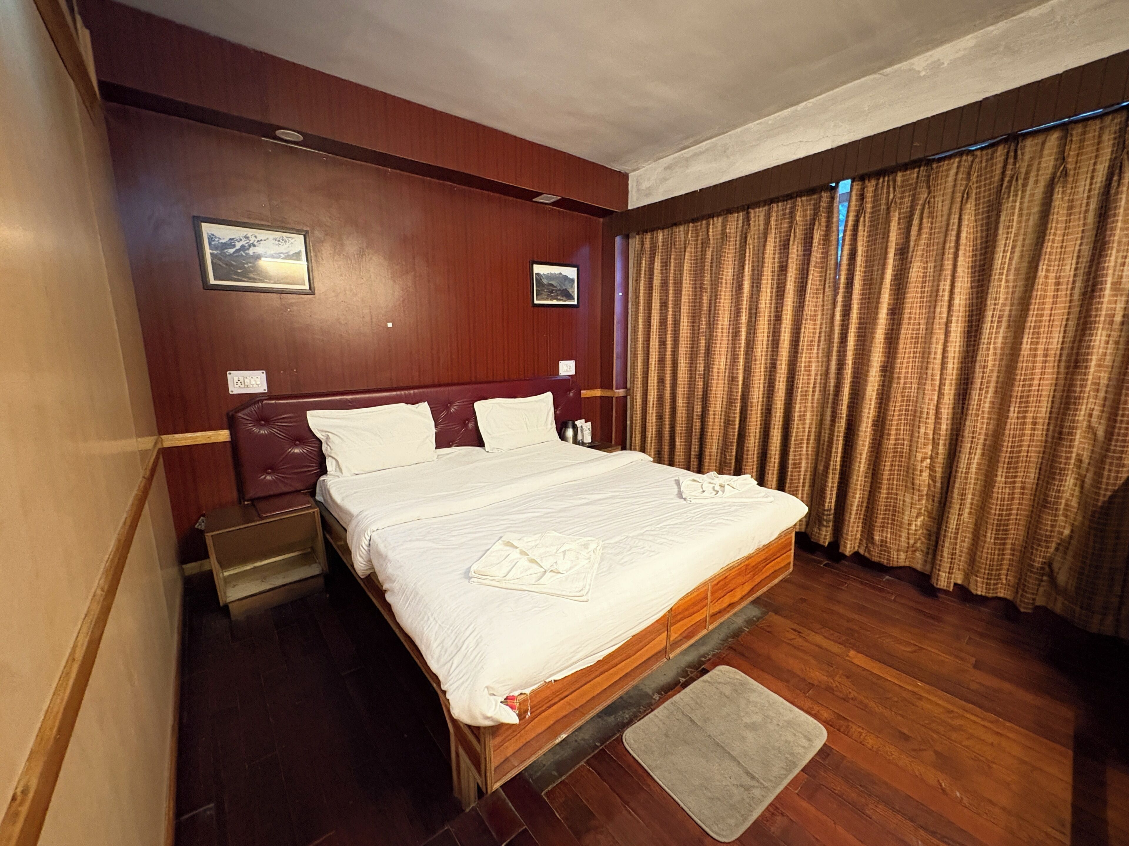 Ein Hotelzimmer mit einem hölzernen Bett, einem Nachttisch, Vorhängen vor dem Fenster und einem an der Wand hängenden Bild.
