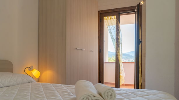 Une chambre d’hôtel avec un lit, une table de chevet avec une lampe, une porte donnant sur un balcon et une vue sur l’extérieur.