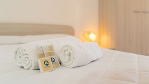 Un lit bien fait, avec des serviettes blanches pliées en rouleau et une carte posée dessus, une armoire en bois et une lampe fixée au mur.