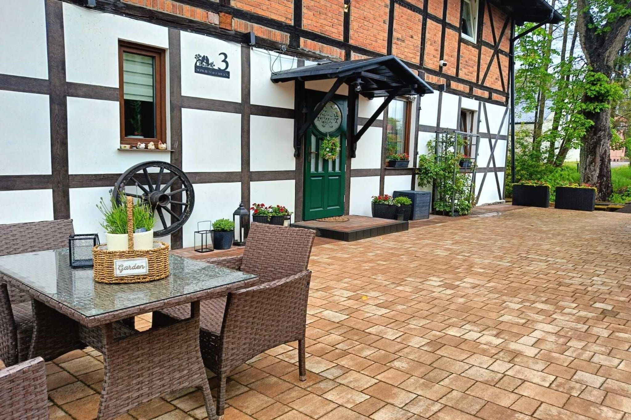 A traditional half-timbered building with a green door, a clock, and a garden area with wicker furniture and potted plants.