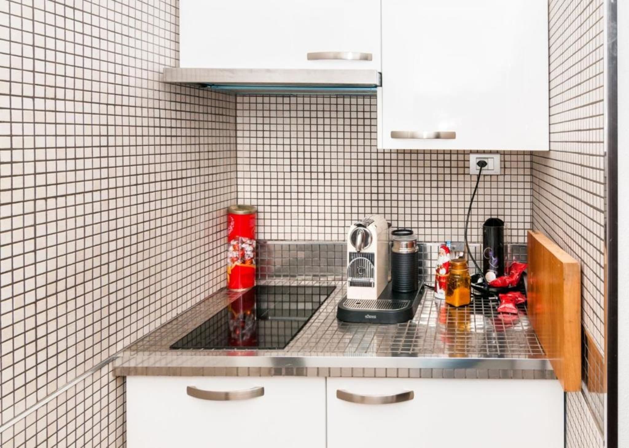 A compact kitchen with a tiled backsplash, a kettle, a coffee maker, and a jar on the counter.