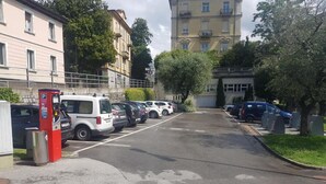 Parkkipaikka, jossa on useita autoja, parkkimittari ja taustalla näkyviä rakennuksia.