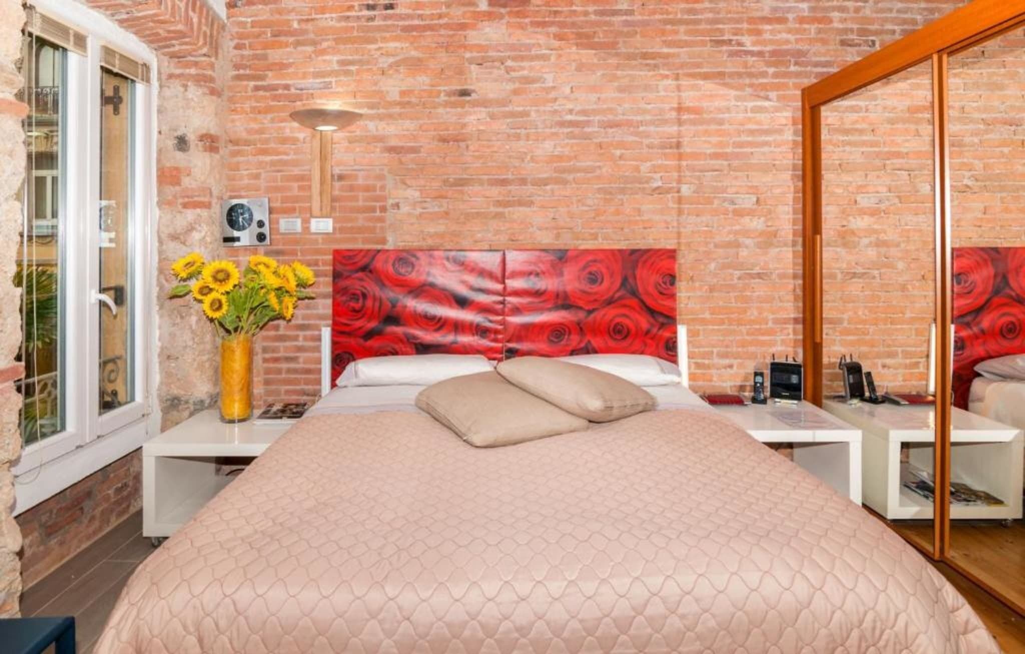 Una habitación con pared de ladrillo, una cama con colcha rosa, un jarrón con girasoles y un cabecero floral rojo.